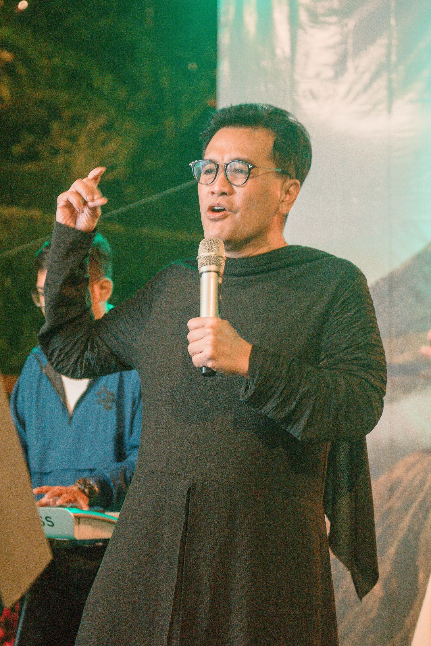 Man in black outfit speaking into microphone with finger raised at an outdoor evening event.