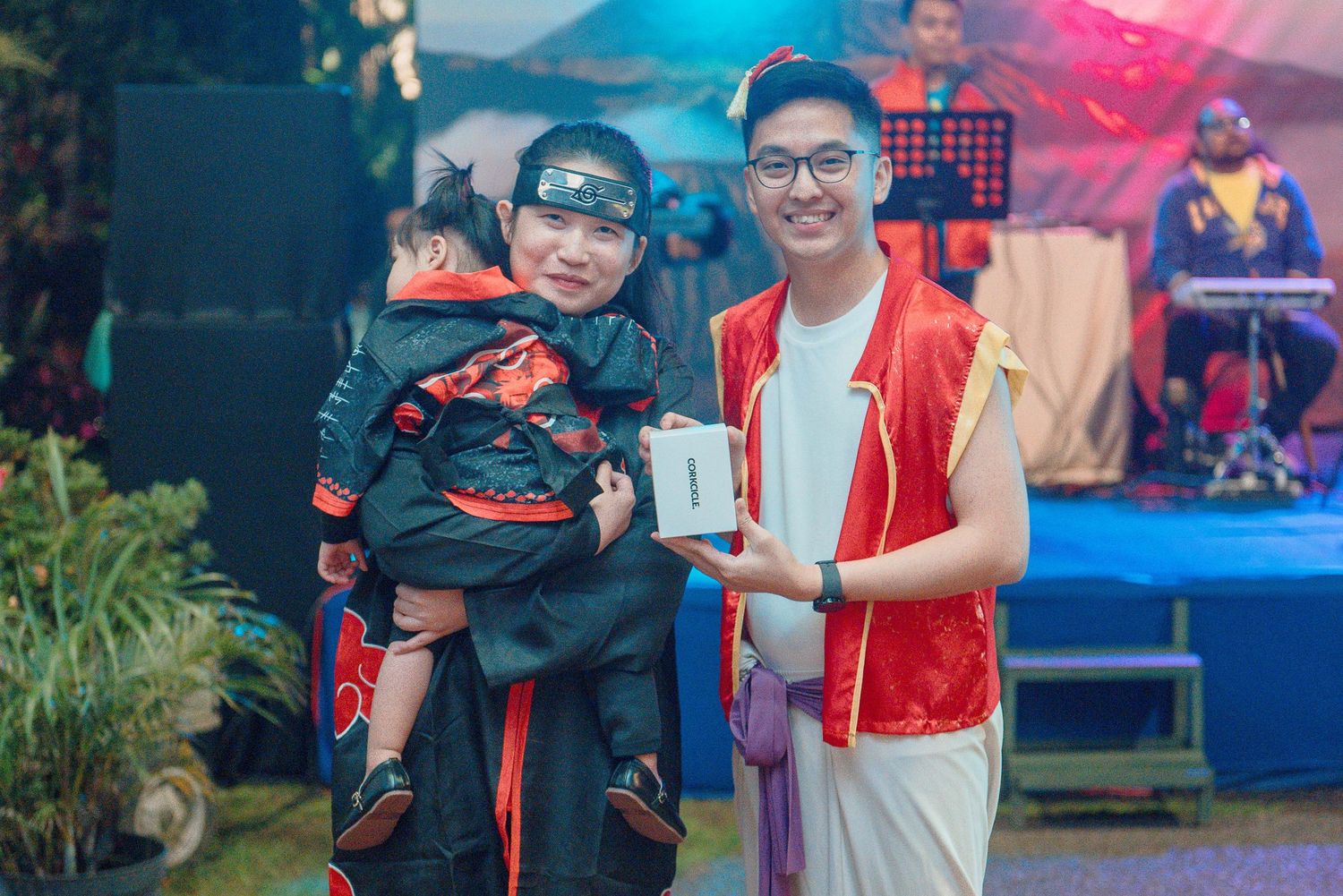 Two people in costumes at an outdoor event, one holding a child dressed as a ninja and a gift box.
