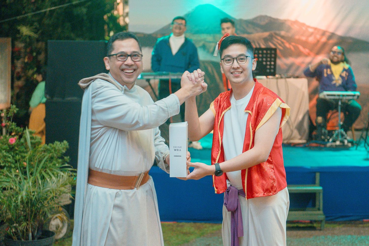 Two men in costumes shaking hands at an outdoor event with a band performing on stage in the background.