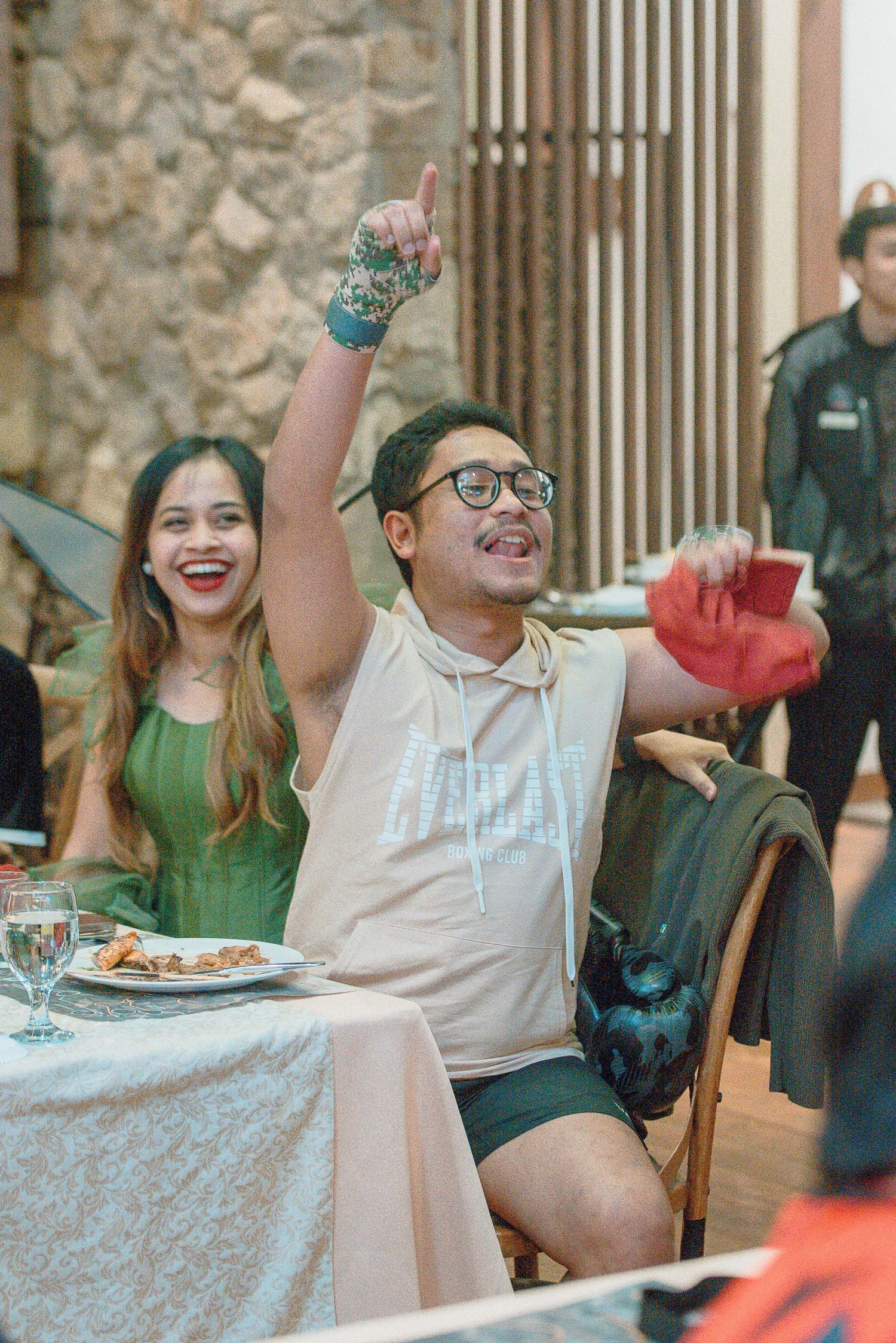 Two people celebrate joyfully at a restaurant table, man raising finger, woman in green dress laughing beside him.