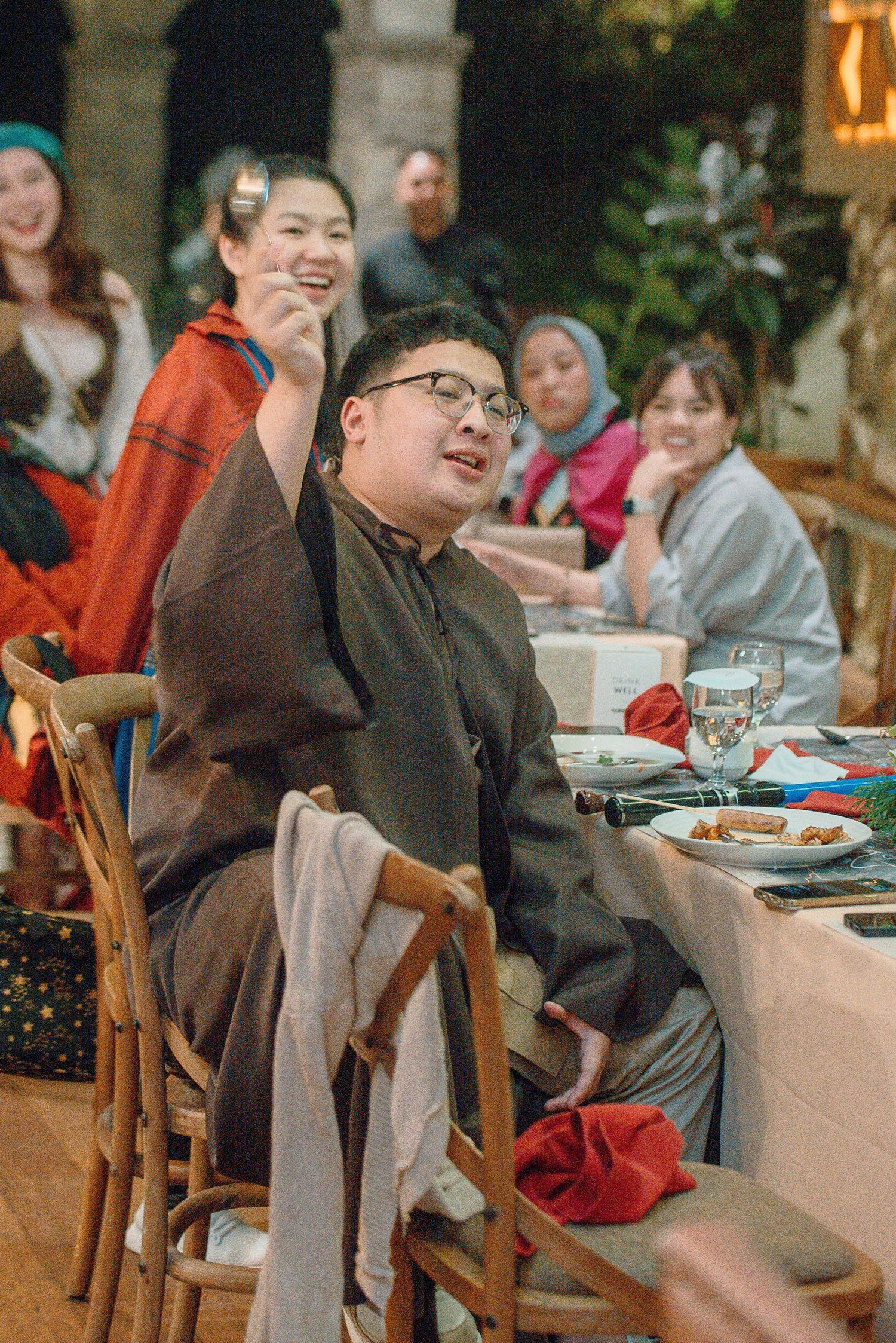 Group of people in costumes enjoying a festive dinner party, with a young man in glasses laughing at a table.