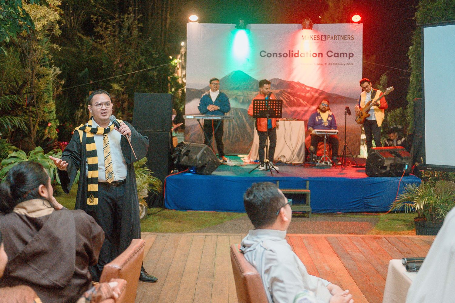 A speaker addresses an audience at a Consolidation Camp event with a live band performing on stage outdoors.