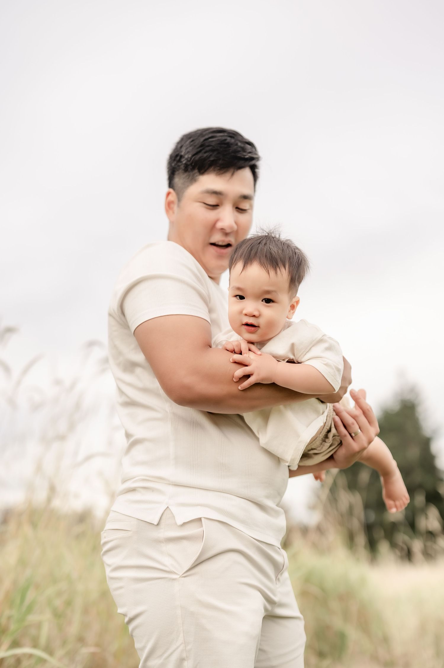 A man in cream outfit holds a baby in a field, both wearing matching neutral tones outdoors.
