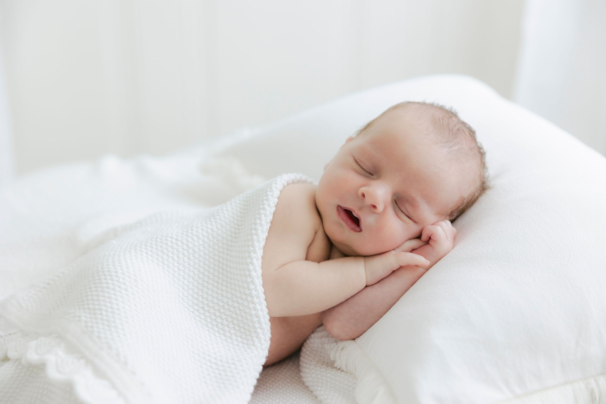 Peaceful newborn baby sleeping on white blanket with hands tucked under cheek in soft natural light.