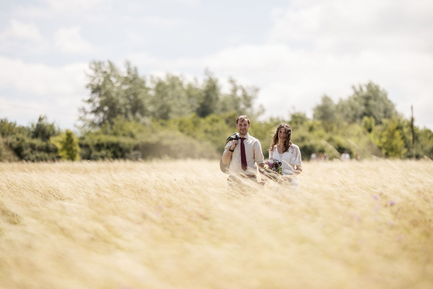 Couple walking through golden wheat field on sunny day with green trees in background.