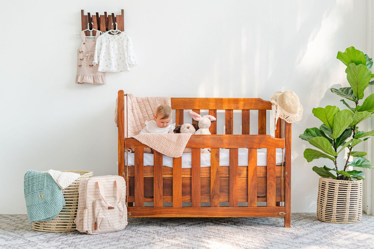 Wooden baby crib with soft bedding, stuffed animals, and a fiddle leaf fig plant in a bright nursery room photographed for a local brand.