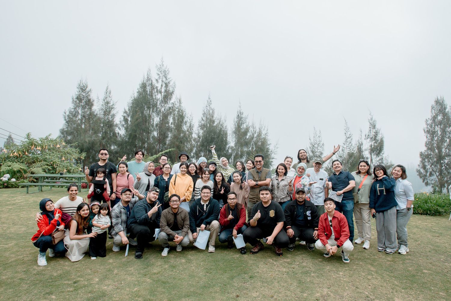 Large group of people posing together outdoors on a grassy hill with pine trees in misty background.