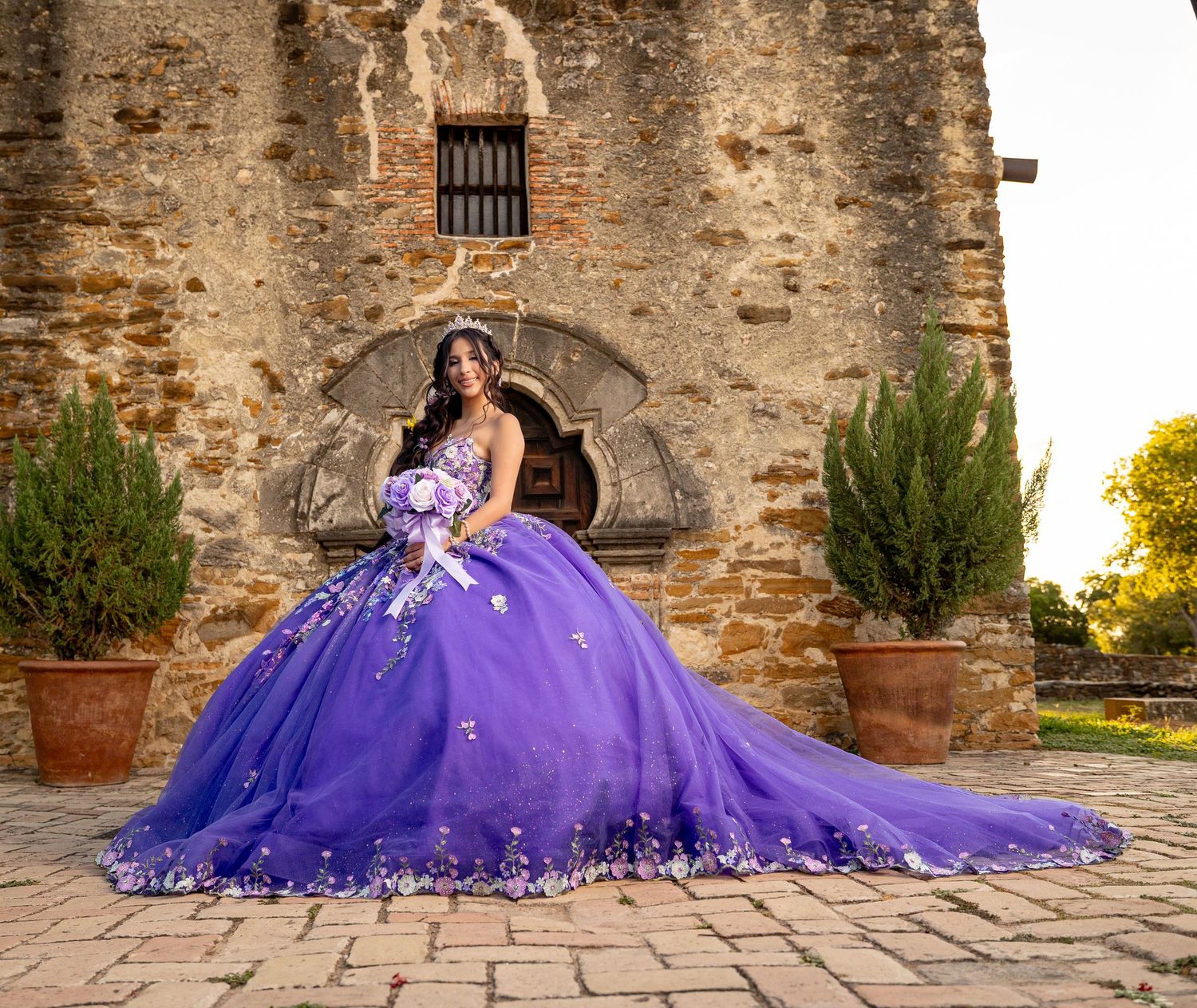 Young woman in purple quinceañera ball gown with floral details poses before historic stone mission building.