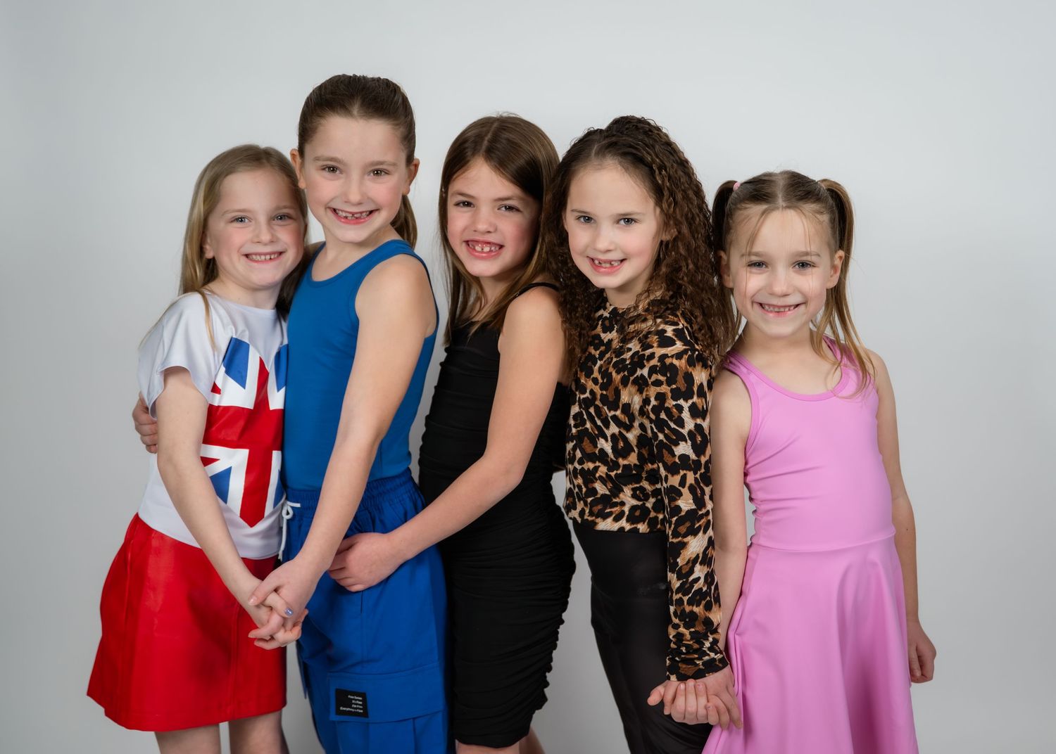Five young girls posing together smiling in colorful outfits against a white background in a professional photo.