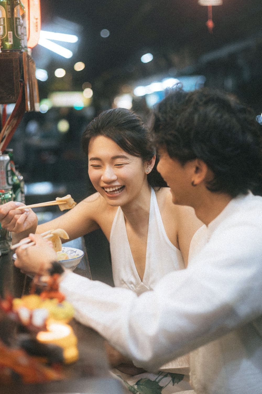Happy Asian couple sharing noodles with chopsticks at a vibrant night market street food stall.