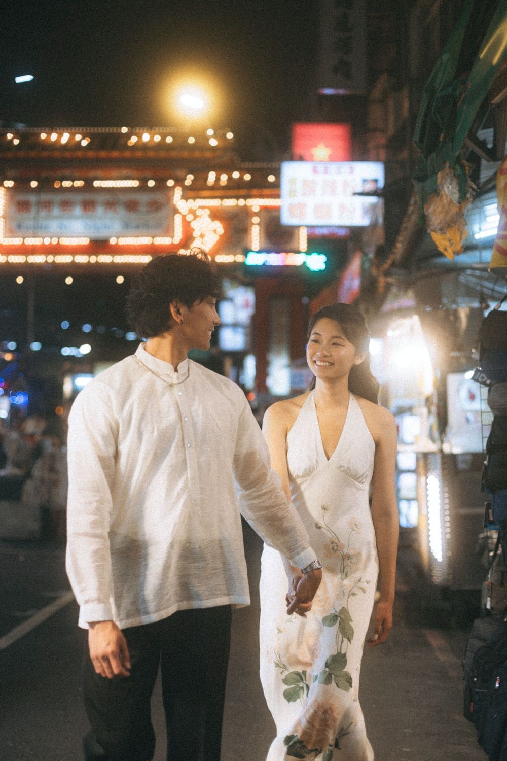 Couple in white outfits holding hands on a vibrant night street with neon signs glowing in the background.