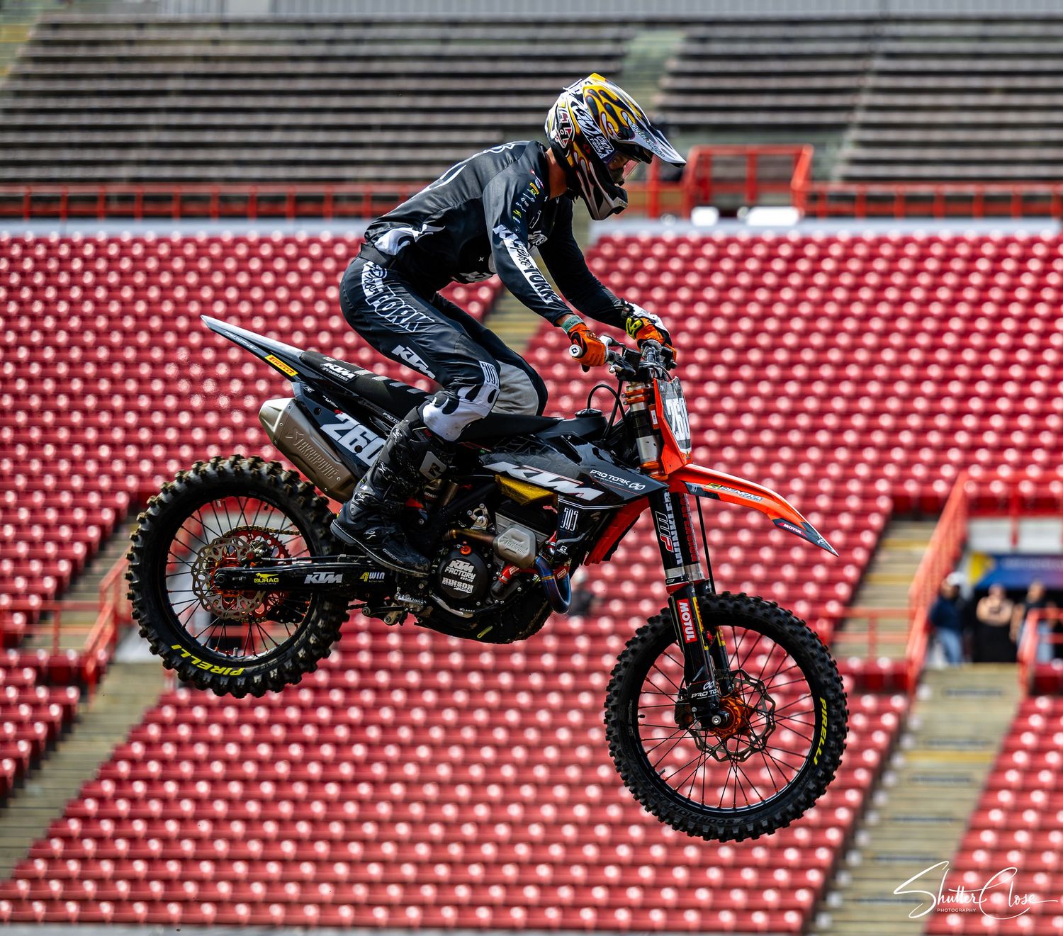 Motocross rider performing a jump on a dirt bike inside a stadium with red empty bleachers in background.