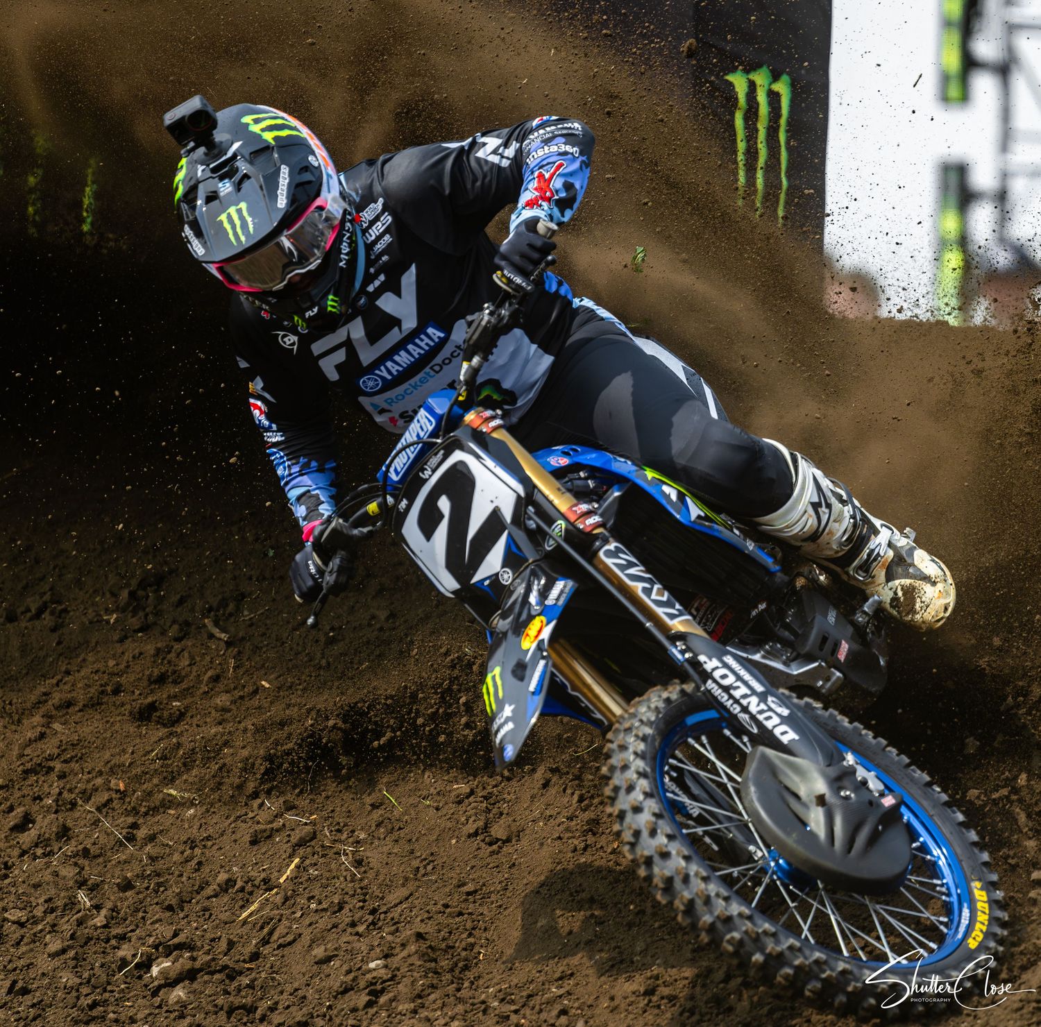 Motocross rider wearing Monster Energy gear races on a blue Yamaha dirt bike through a muddy track.