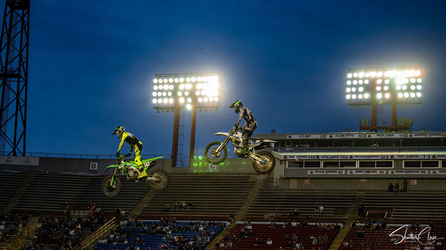 Two motocross riders perform aerial jumps at a night stadium event under bright floodlights.