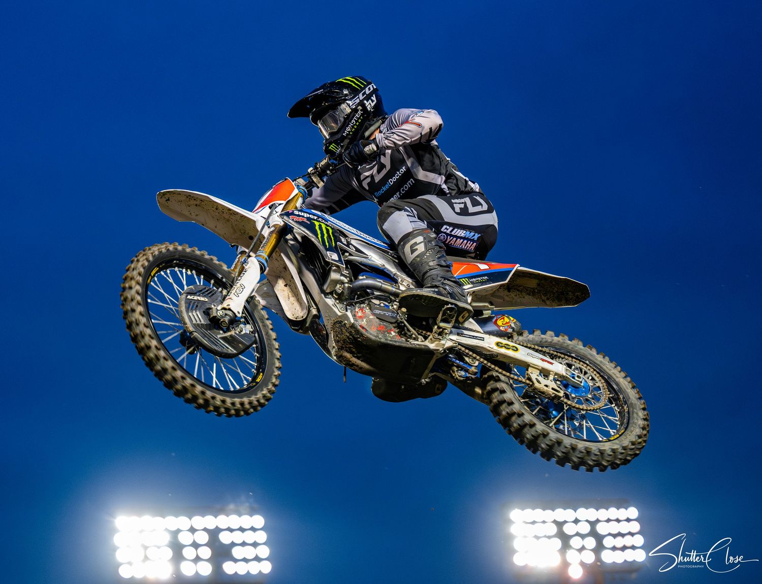 Motocross rider performs a high jump on a dirt bike under bright stadium lights against a deep blue night sky.