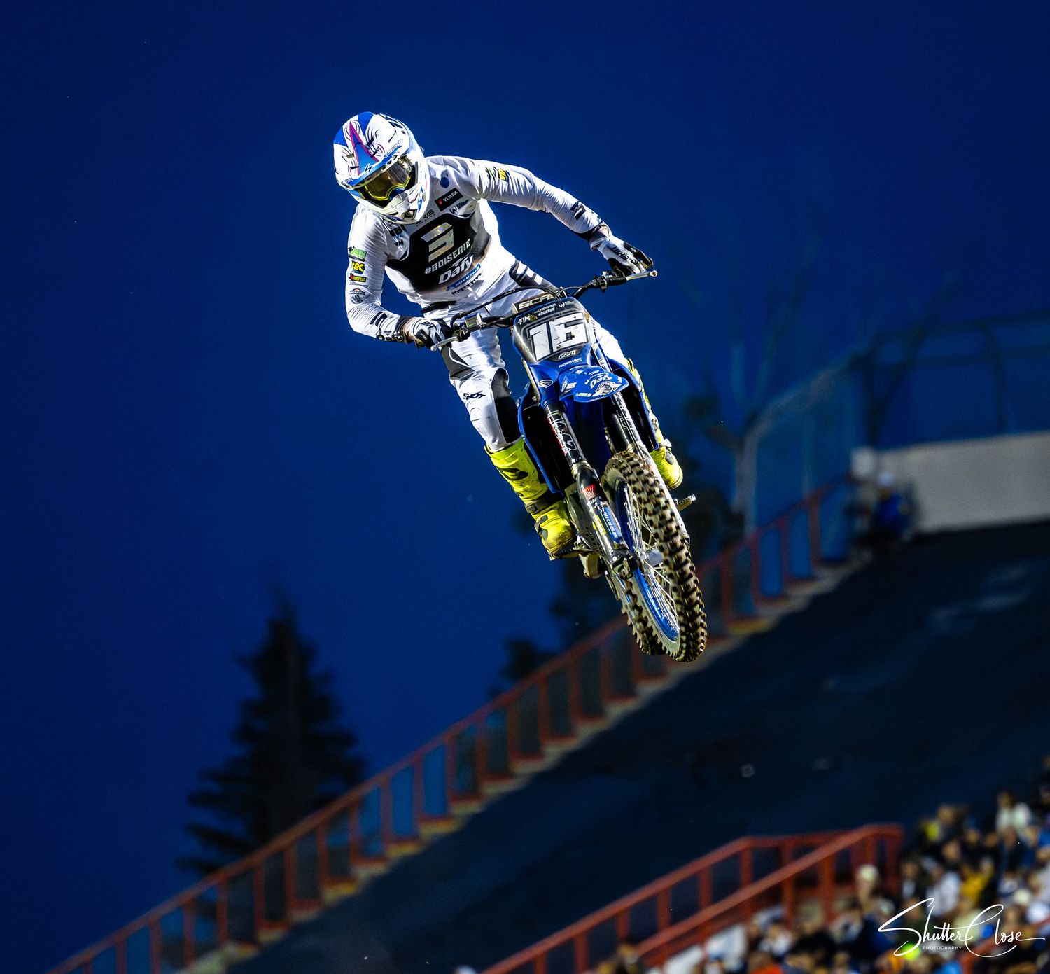 Motocross rider soaring through the air on a yellow dirt bike during a nighttime supercross event.