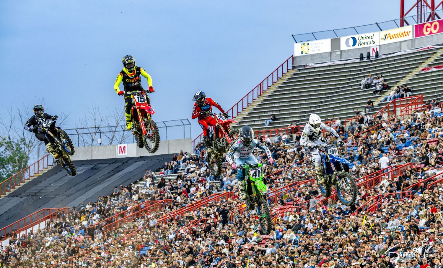 Motocross riders compete on a dirt track hill before a packed stadium crowd under clear blue skies.