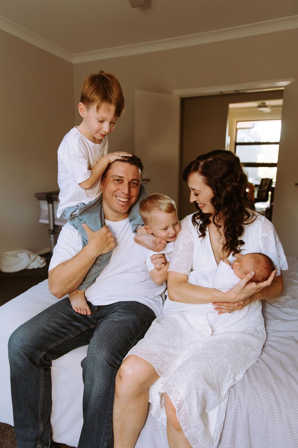 Happy family of five sitting on bed, parents with three children including newborn baby, smiling together at home.