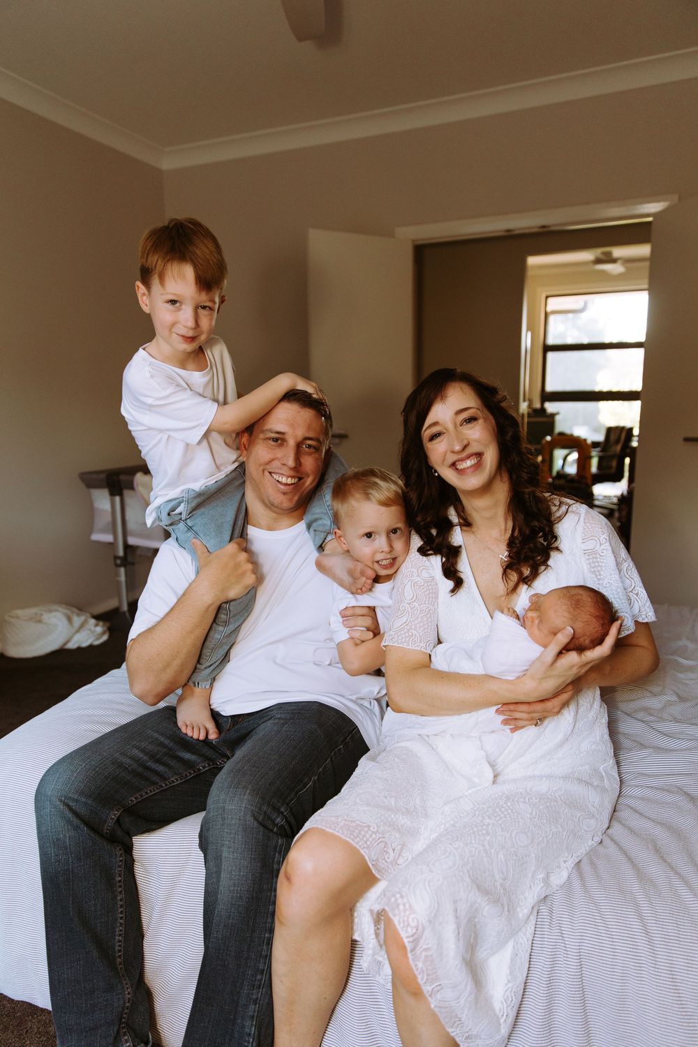 Happy family of five posing on a bed, parents smiling with two young boys and a newborn baby in a cozy bedroom.
