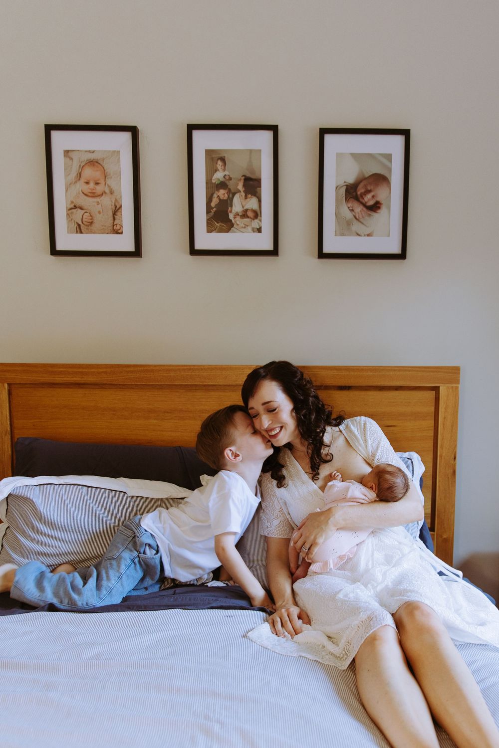 Mother in white dress sits on bed holding newborn while young child leans in to kiss her cheek, framed photos above.