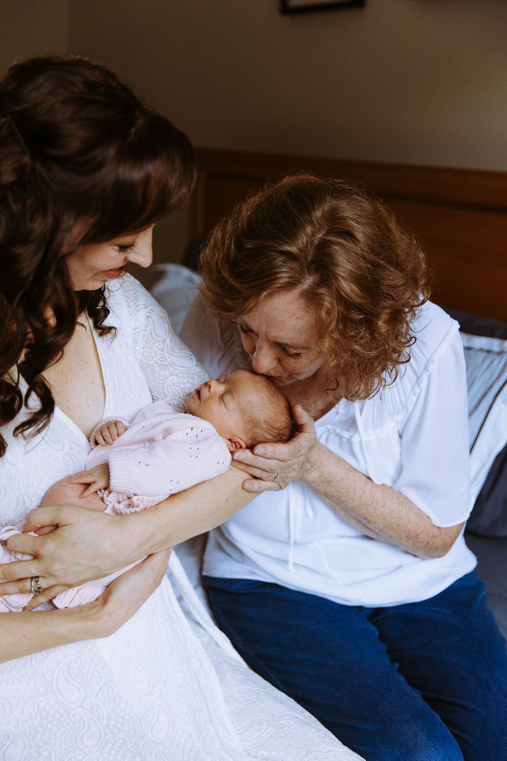 A mother in white holds her newborn in pink while an older woman lovingly kisses the baby on a bed.
