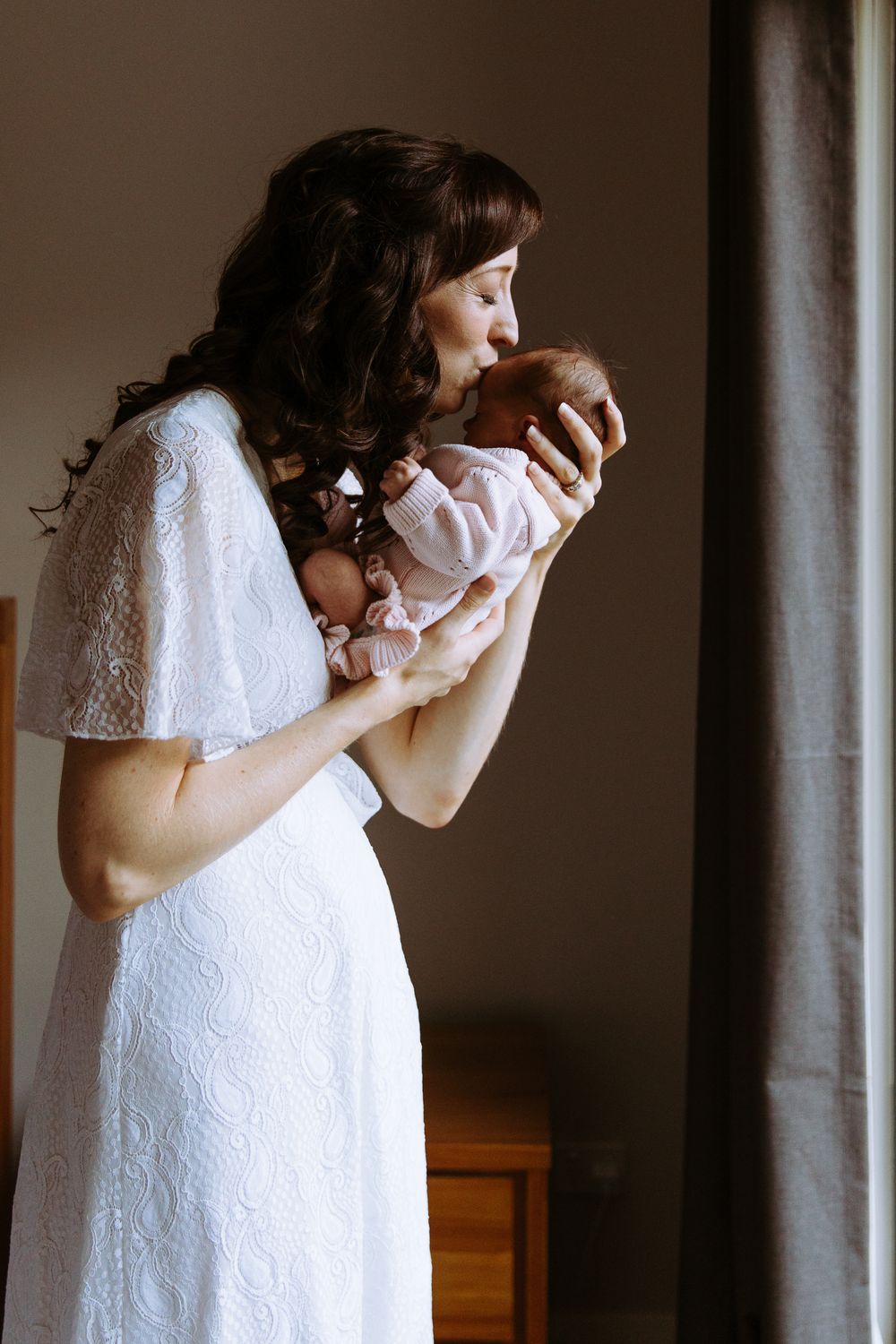 A mother in a white lace dress tenderly kisses her newborn baby while standing by a window.