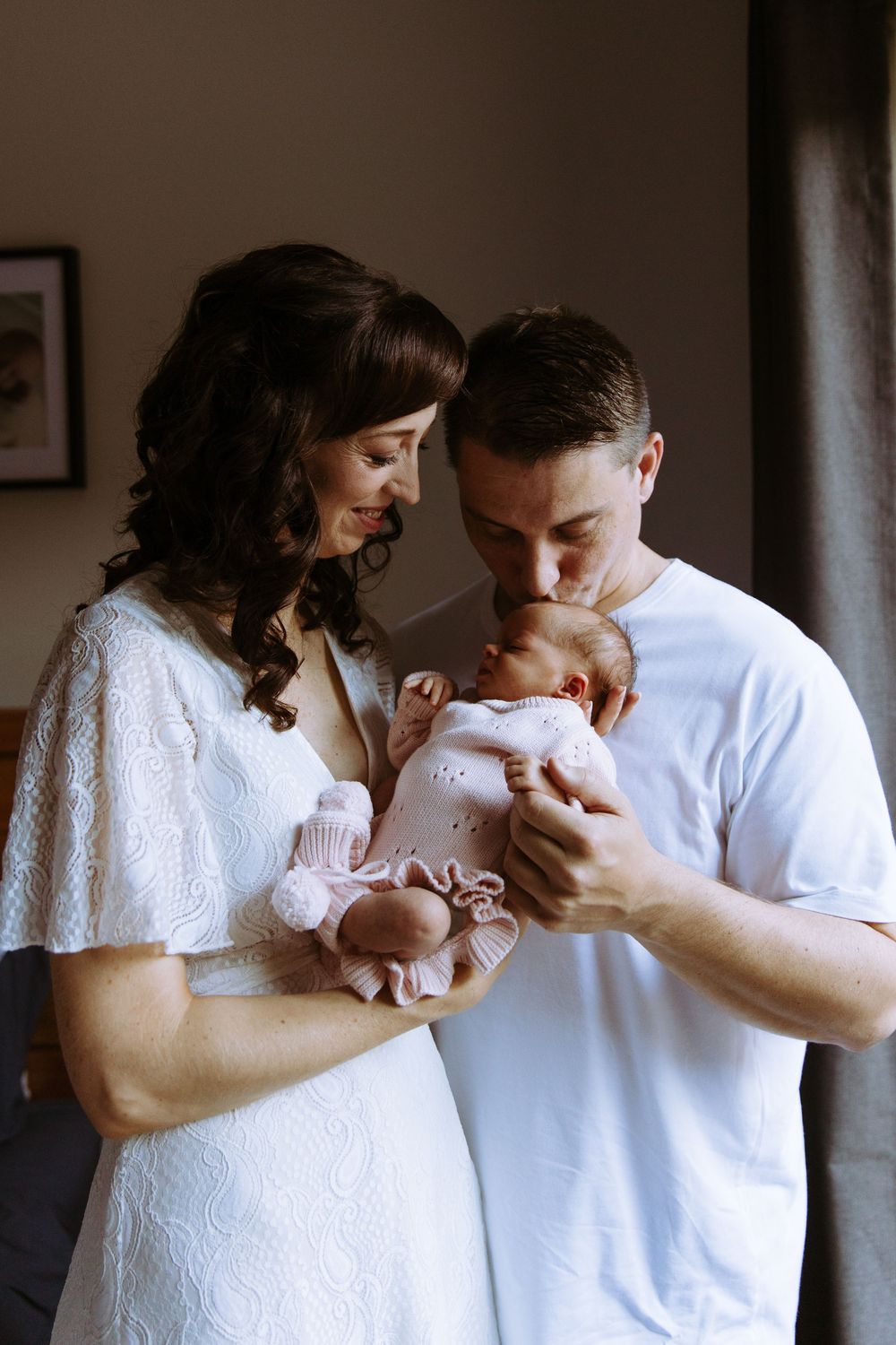 New parents in white outfits tenderly holding and kissing their sleeping newborn baby dressed in pink.