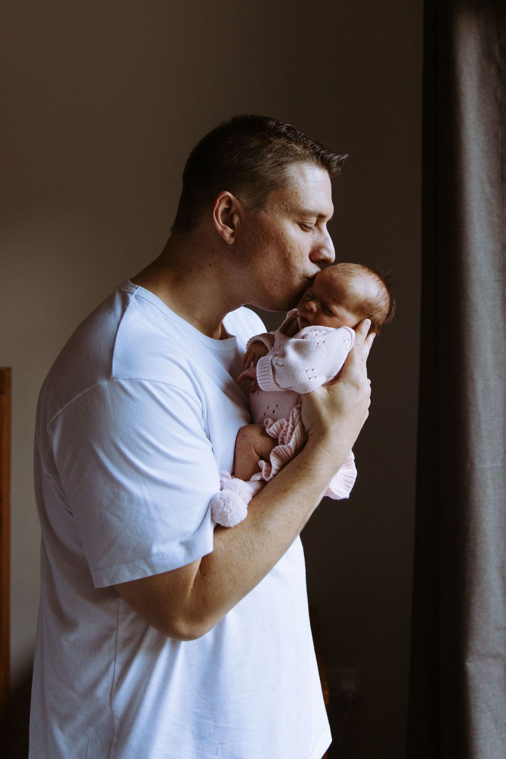 A father tenderly kisses his newborn baby dressed in pink while standing by a window.