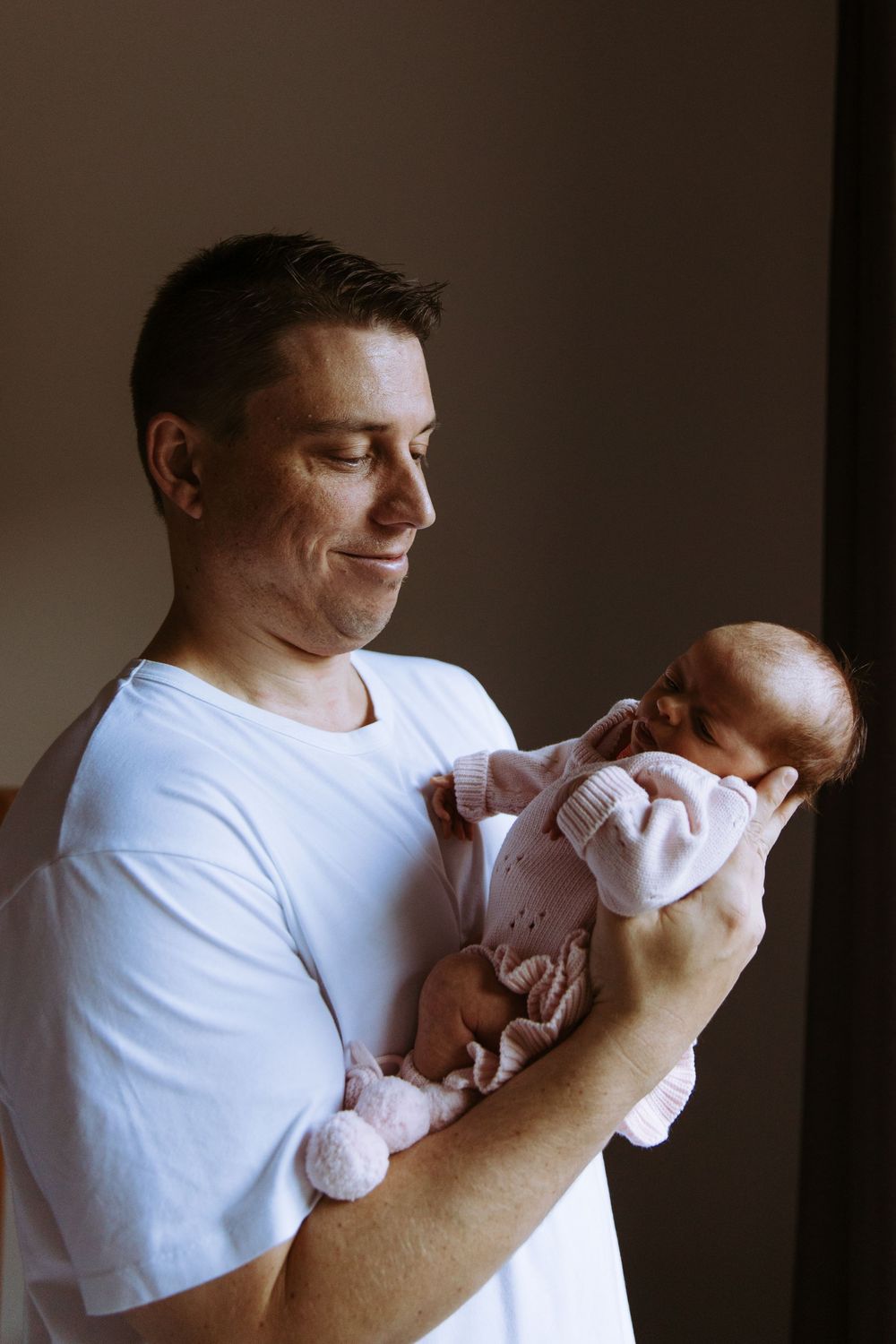 A smiling father in a white t-shirt gently holds his sleeping newborn baby dressed in pink.