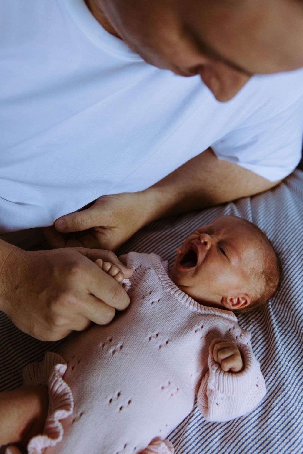 Father in white shirt tenderly holds crying newborn baby dressed in pink knit outfit on striped blanket.