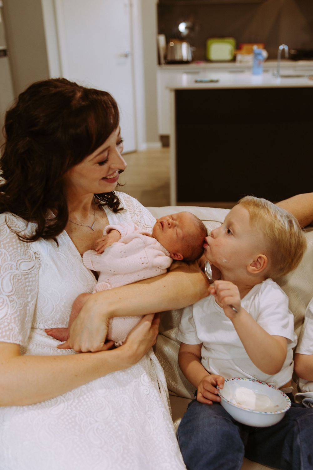A smiling mother in a white dress holds her newborn baby while her toddler son offers a spoonful of food.