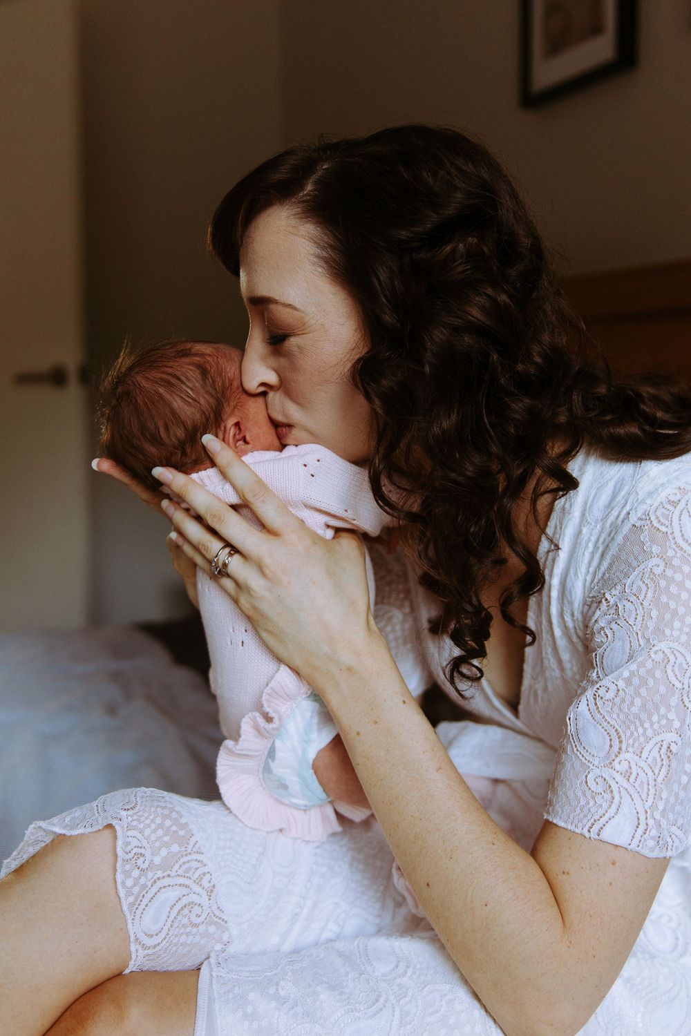 Tender moment of a curly-haired mother in a white lace dress kissing her newborn baby while seated on a bed.