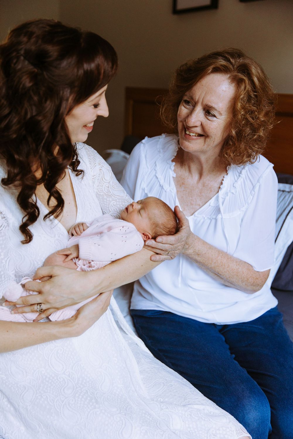 Two women in white tops admire a newborn baby, sharing a tender three-generation family moment indoors.
