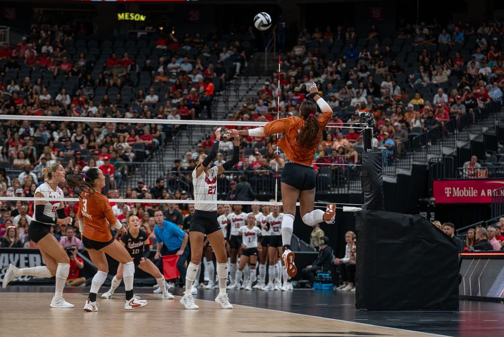 College volleyball players in orange uniforms compete at net during an indoor arena match under bright lights.