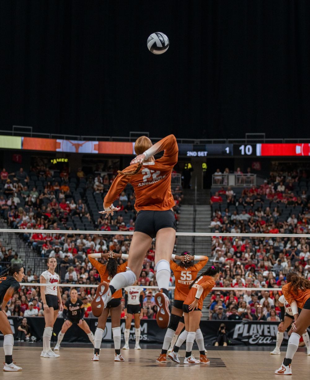 Texas Longhorns volleyball player jumps to spike the ball during a competitive indoor match at a packed arena.