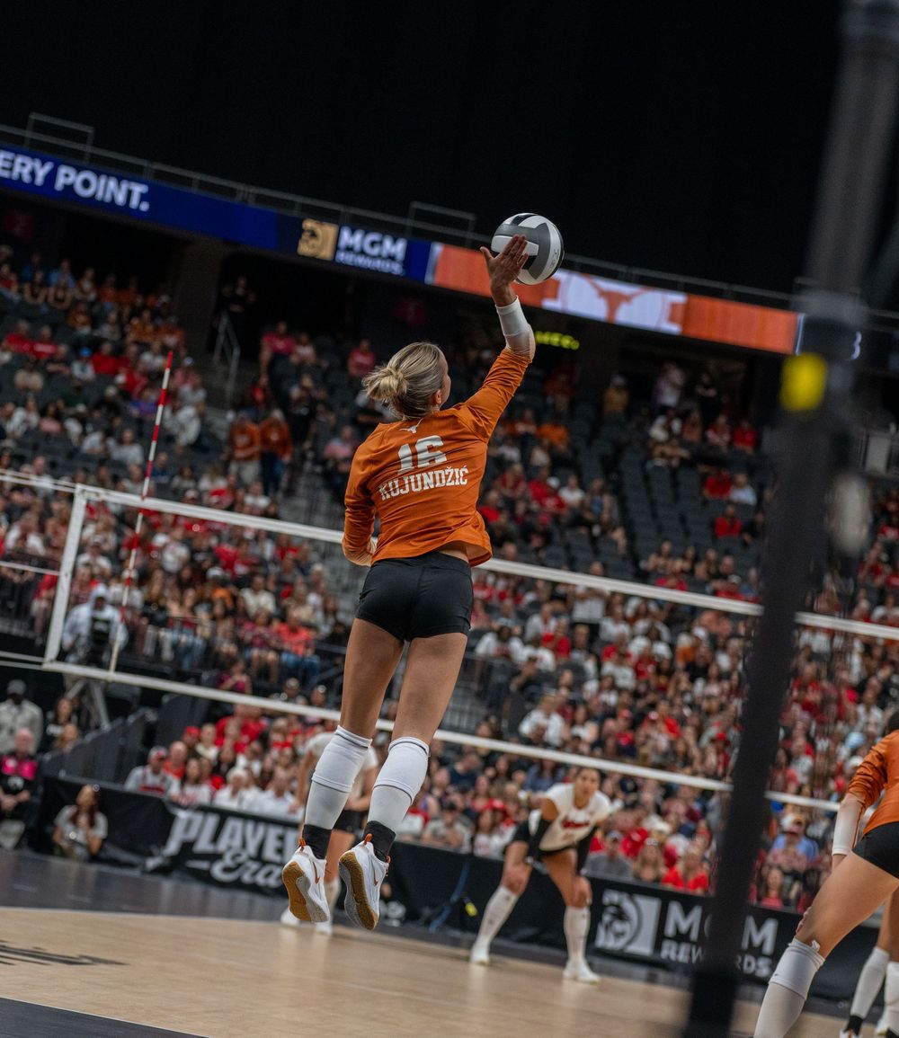 College volleyball player in orange jersey #15 jumps to spike the ball during a packed arena match.