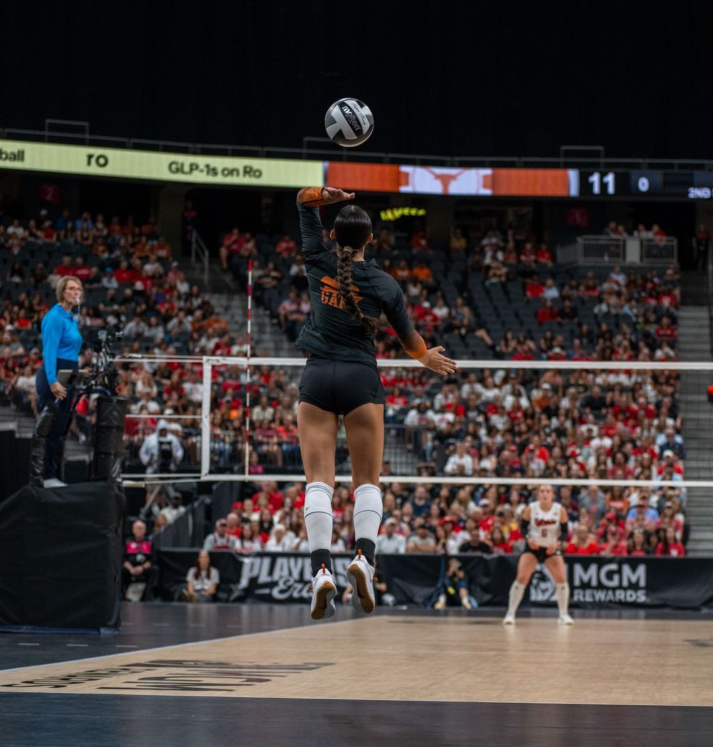 Volleyball player jumping to spike the ball during a competitive indoor arena match with packed crowd.