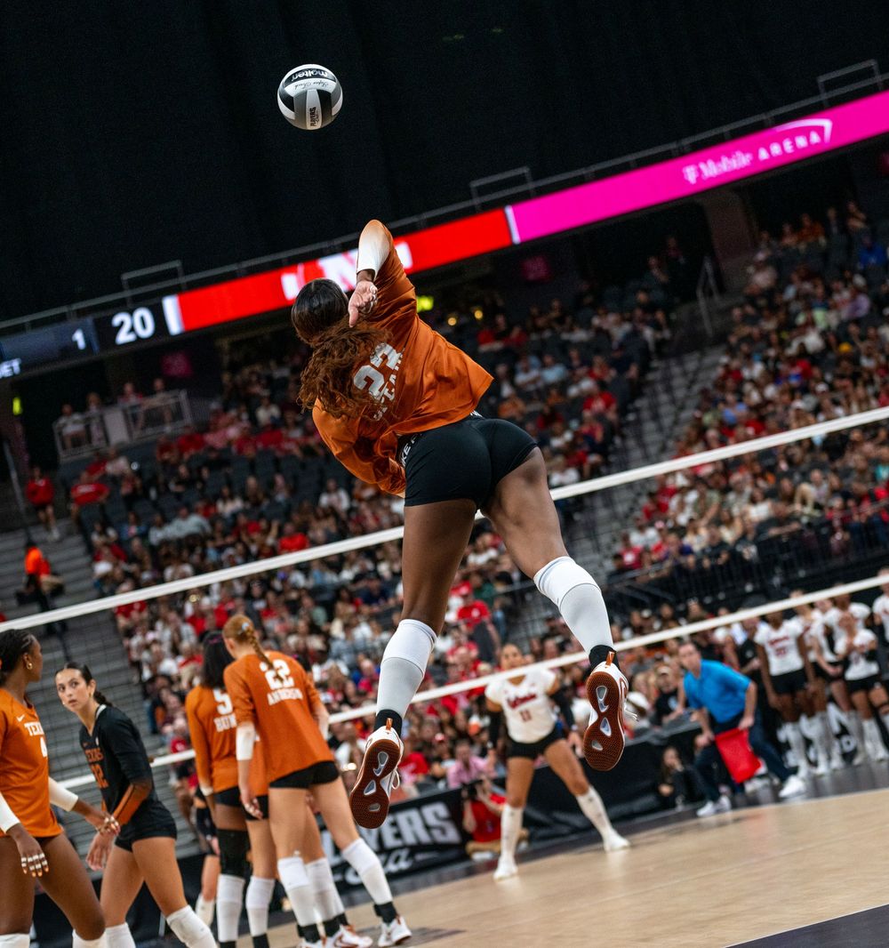Female volleyball player in orange jersey jumps to spike ball over net during competitive indoor match.