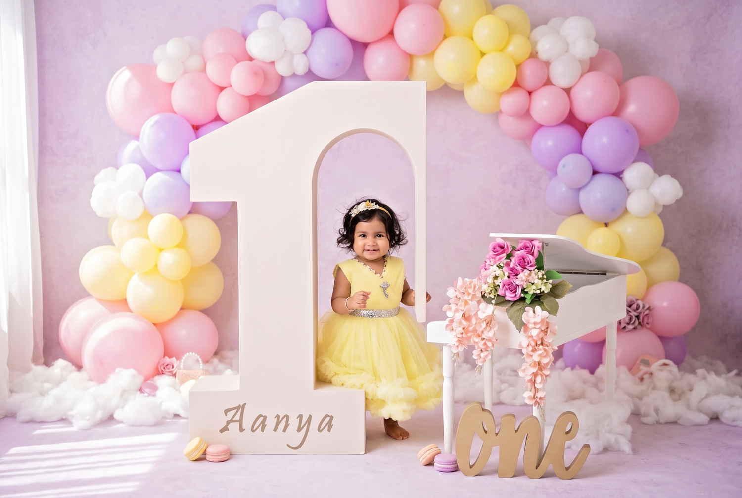 Baby girl in pink tutu sits inside a large number 1 prop decorated with balloons and lights for her first birthday.