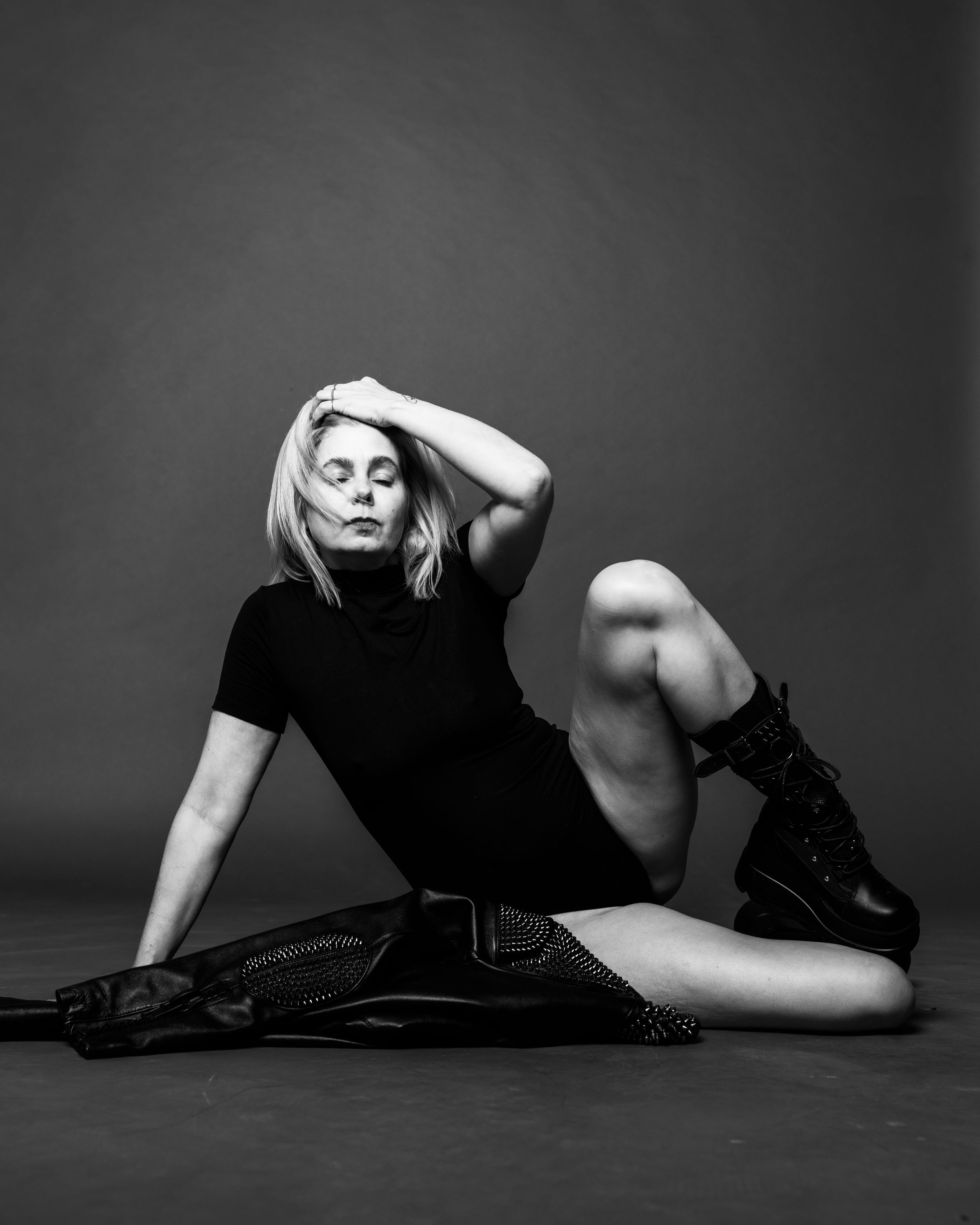 Black and white photo of a blonde woman in a black dress posing on the floor with her hand behind her head.