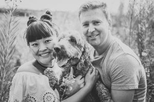 A happy couple poses with their scruffy terrier dog outdoors in black and white.