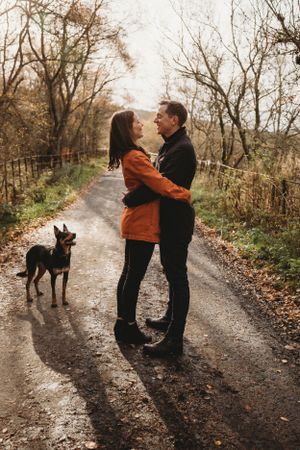 Couple embracing on an autumn path while their dog watches nearby.