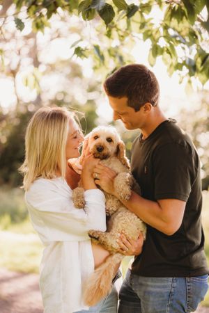 Couple holding their goldendoodle puppy outdoors in warm sunlight under trees.