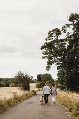 A couple walking their dog down a country path surrounded by tall grass and trees on a cloudy day.