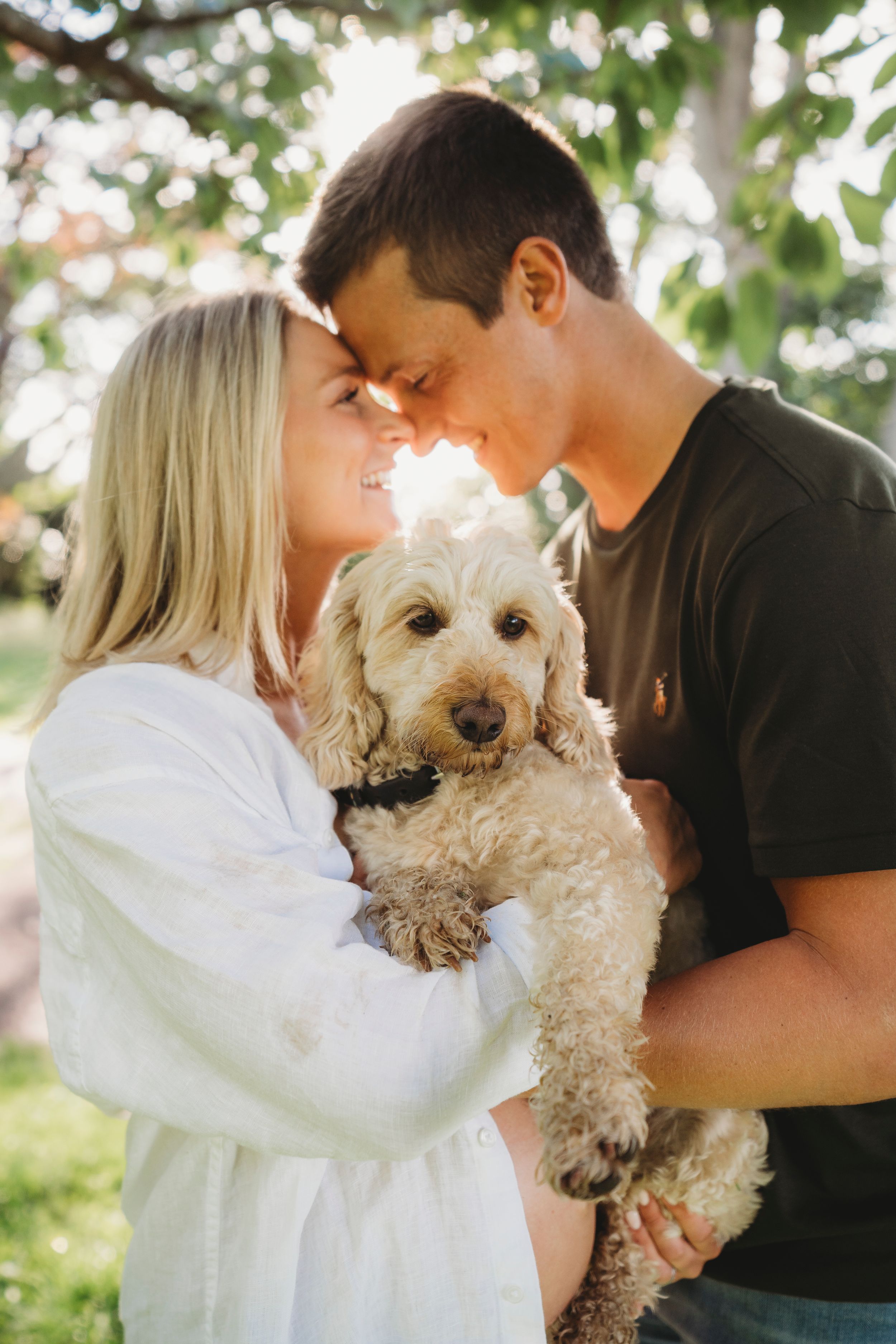A tender moment between a couple holding their fluffy goldendoodle dog in a sunlit garden setting.