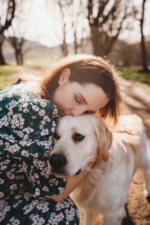 Person in floral dress hugging golden retriever in sunlit outdoor setting.