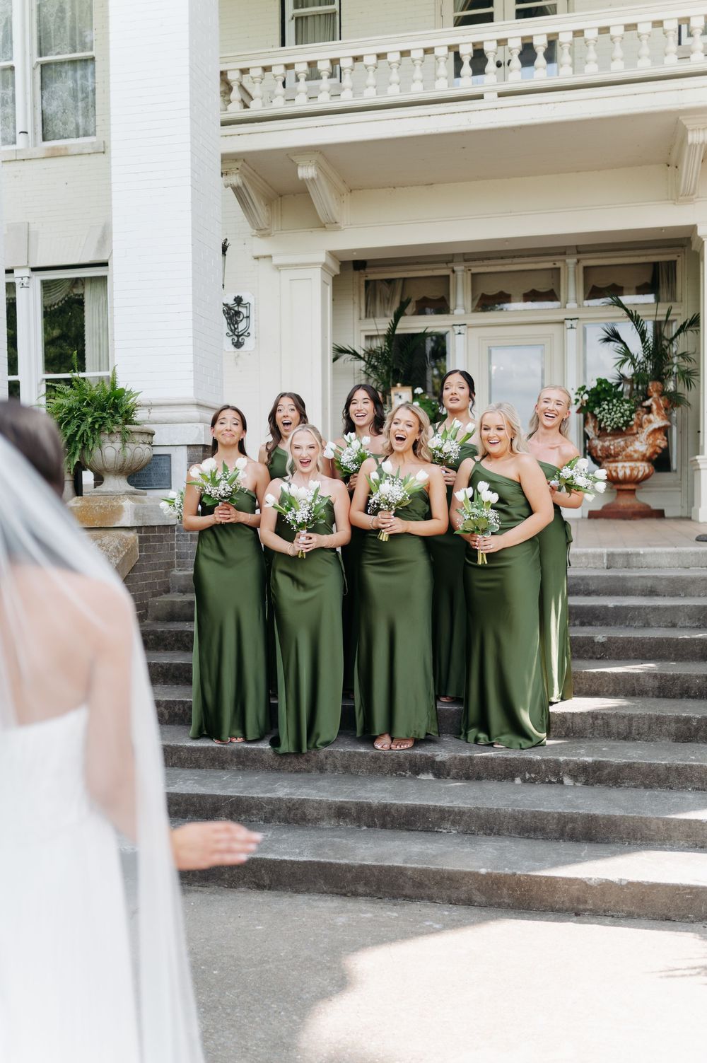 Bridesmaids in green gowns holding white bouquets react with joy seeing the bride on mansion steps.