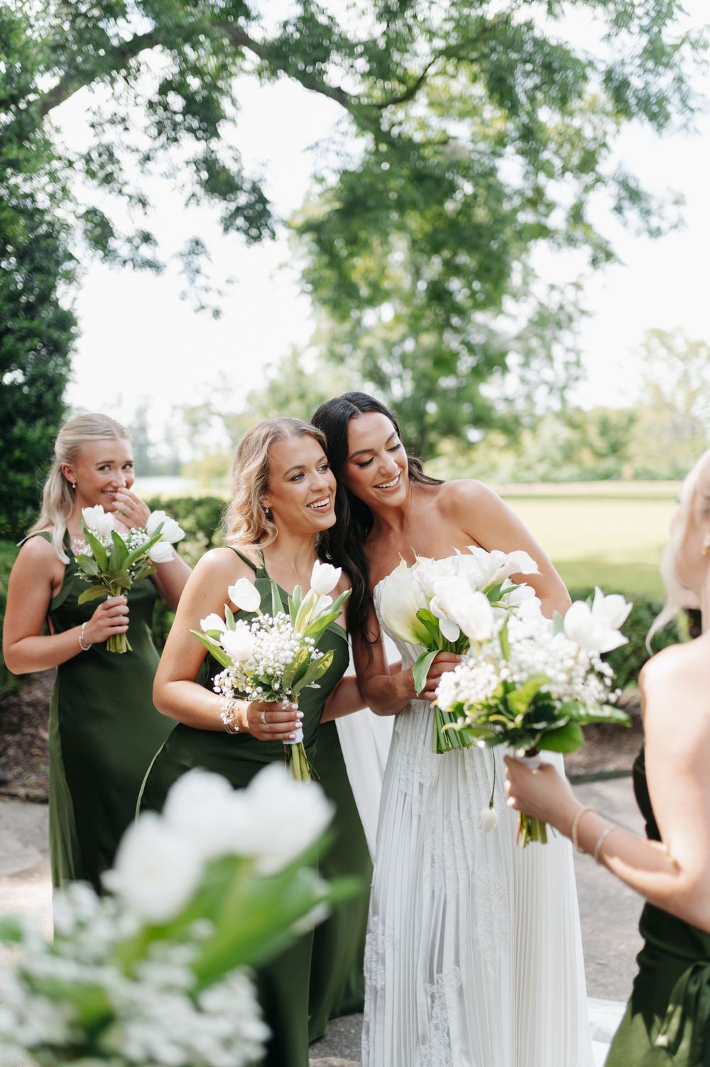 Bride in white gown with bridesmaids in olive green dresses holding white bouquets outdoors at a wedding celebration.