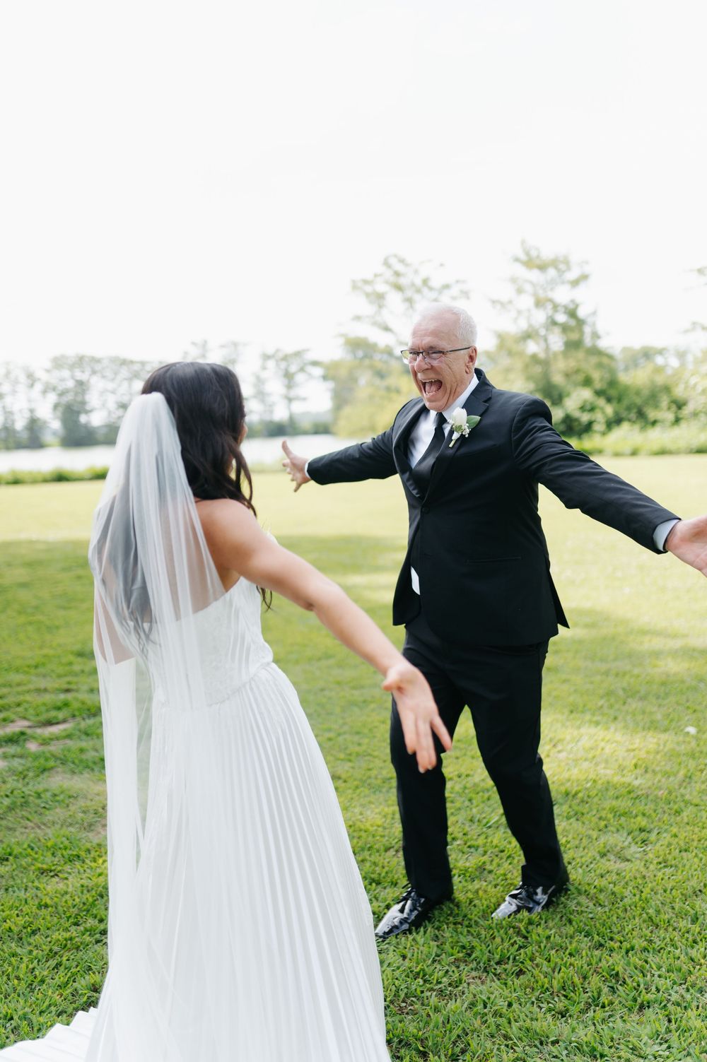 Bride in white pleated gown and veil shares an emotional first look moment with older man in black suit outdoors.
