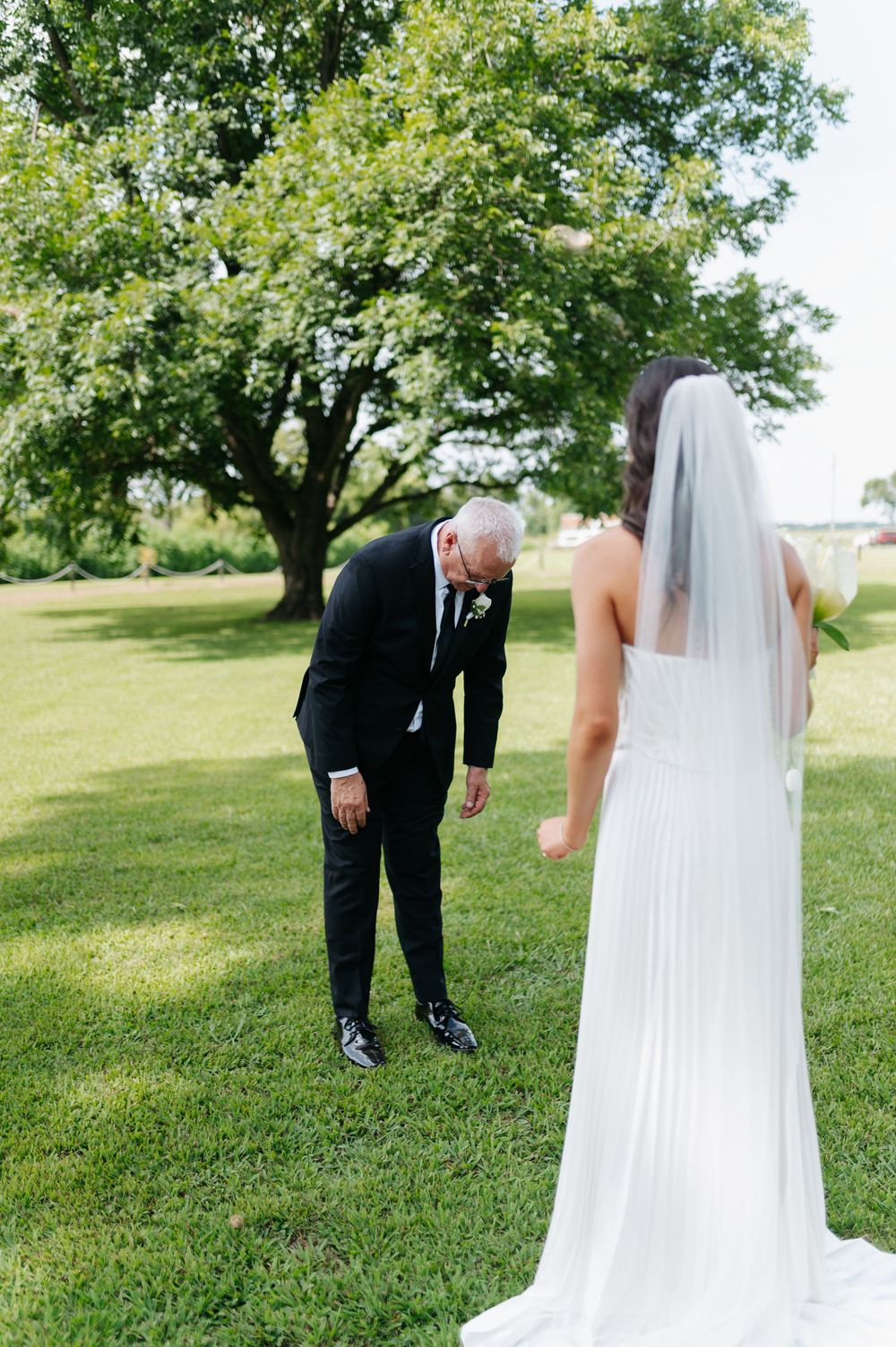 Emotional father bows head seeing bride in white pleated wedding gown and veil for first look on green lawn.